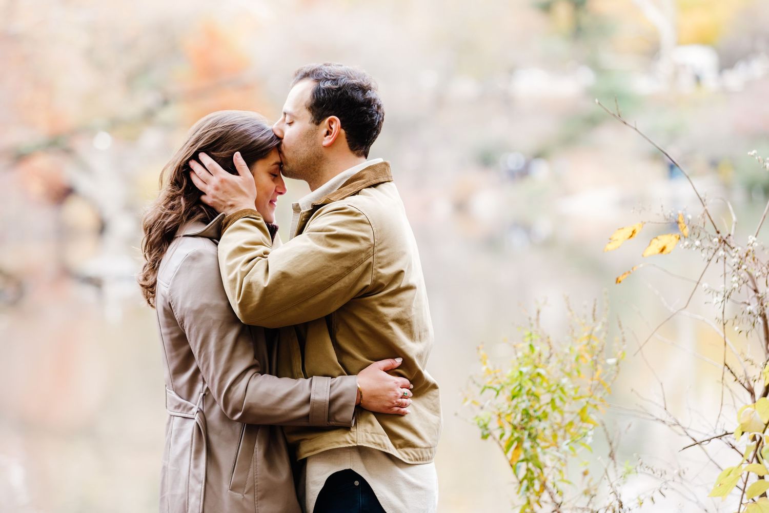 Romantic couple embracing and touching foreheads near a tranquil autumn waterside setting.
