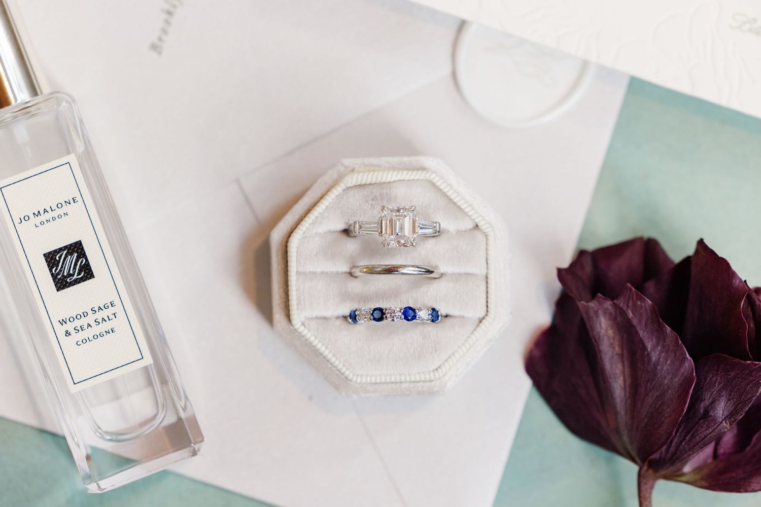 Wedding rings in velvet box beside Jo Malone perfume bottle and dark purple tulip on white surface.