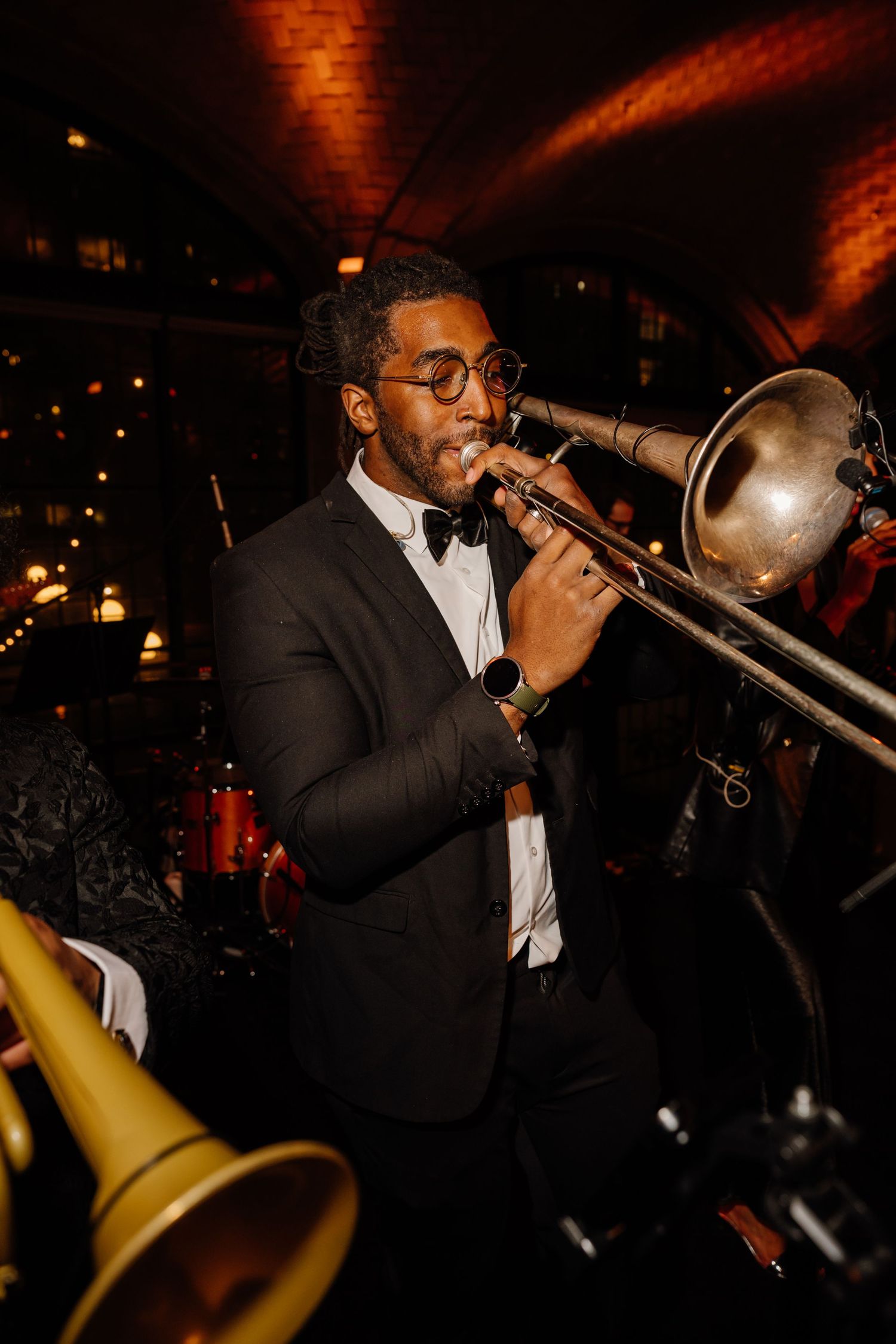 Talented musician in tuxedo plays trombone at a lively wedding reception in a warmly lit venue.