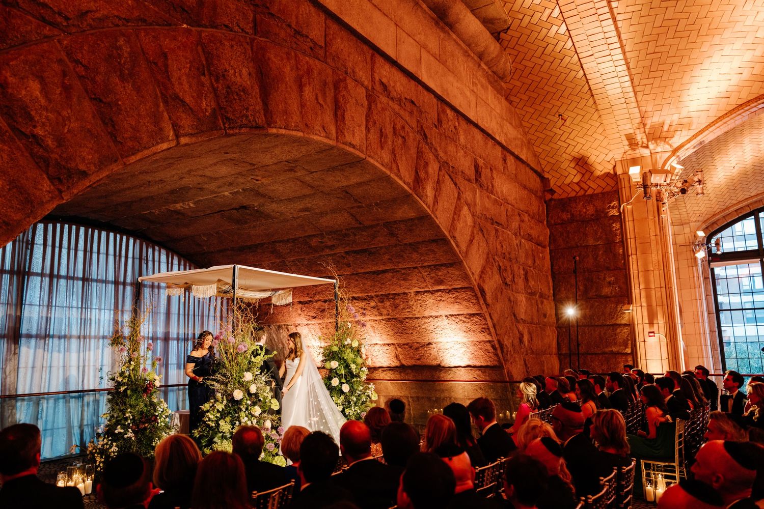 Bride and groom stand under chuppah during elegant wedding ceremony in grand vaulted stone venue with warm amber lighting.