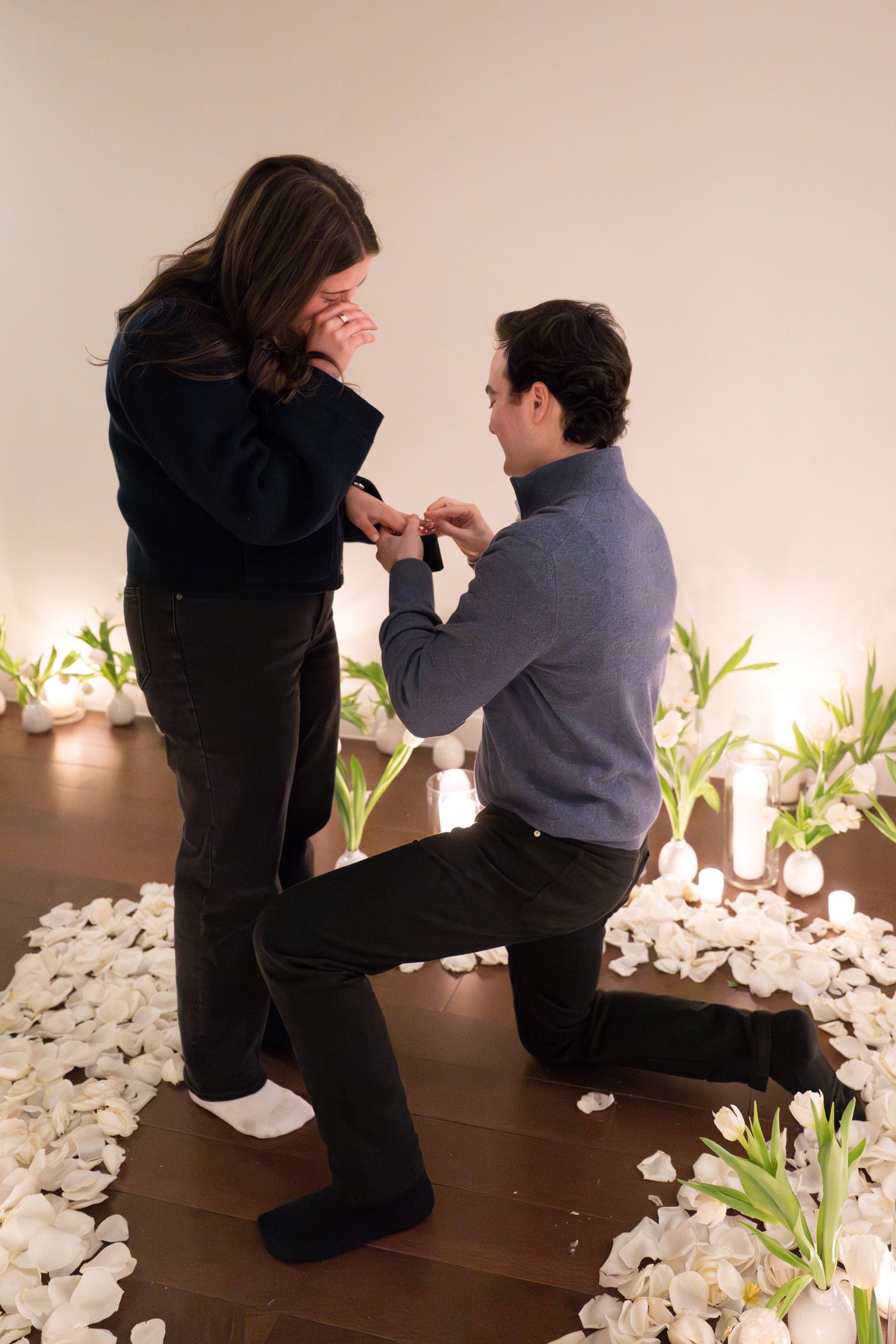 Man on one knee placing an engagement ring on his emotional girlfriends finger amid white tulips and rose petals.