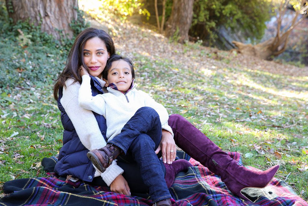 A woman and young child sit together on a plaid blanket outdoors, smiling warmly in a park setting.
