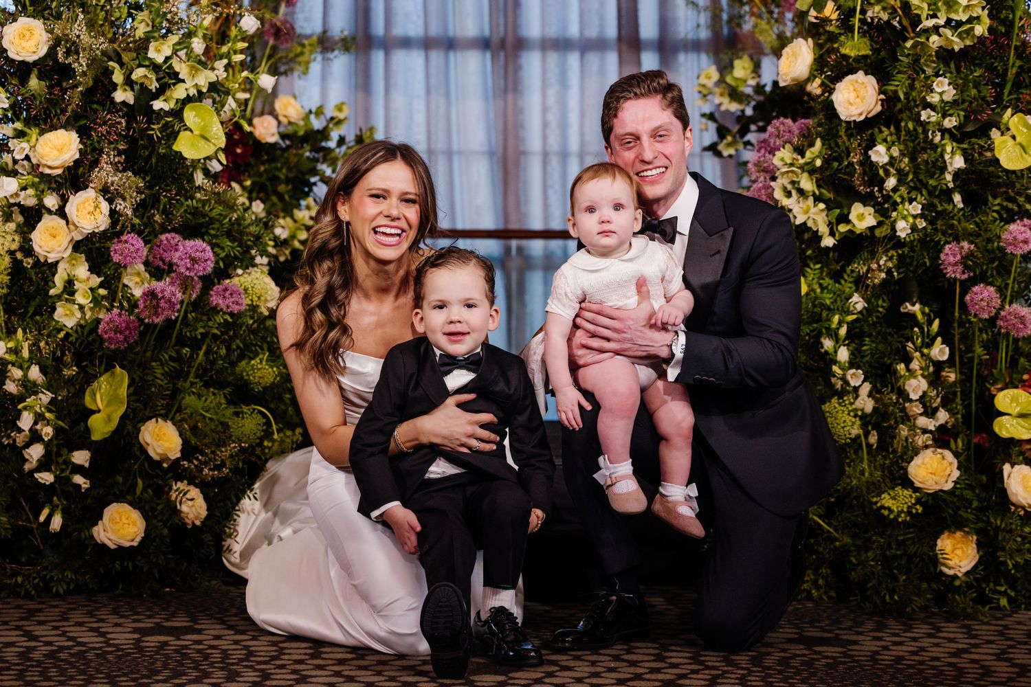 Newlywed couple kneeling with two young children in formal wear before floral wedding backdrop.