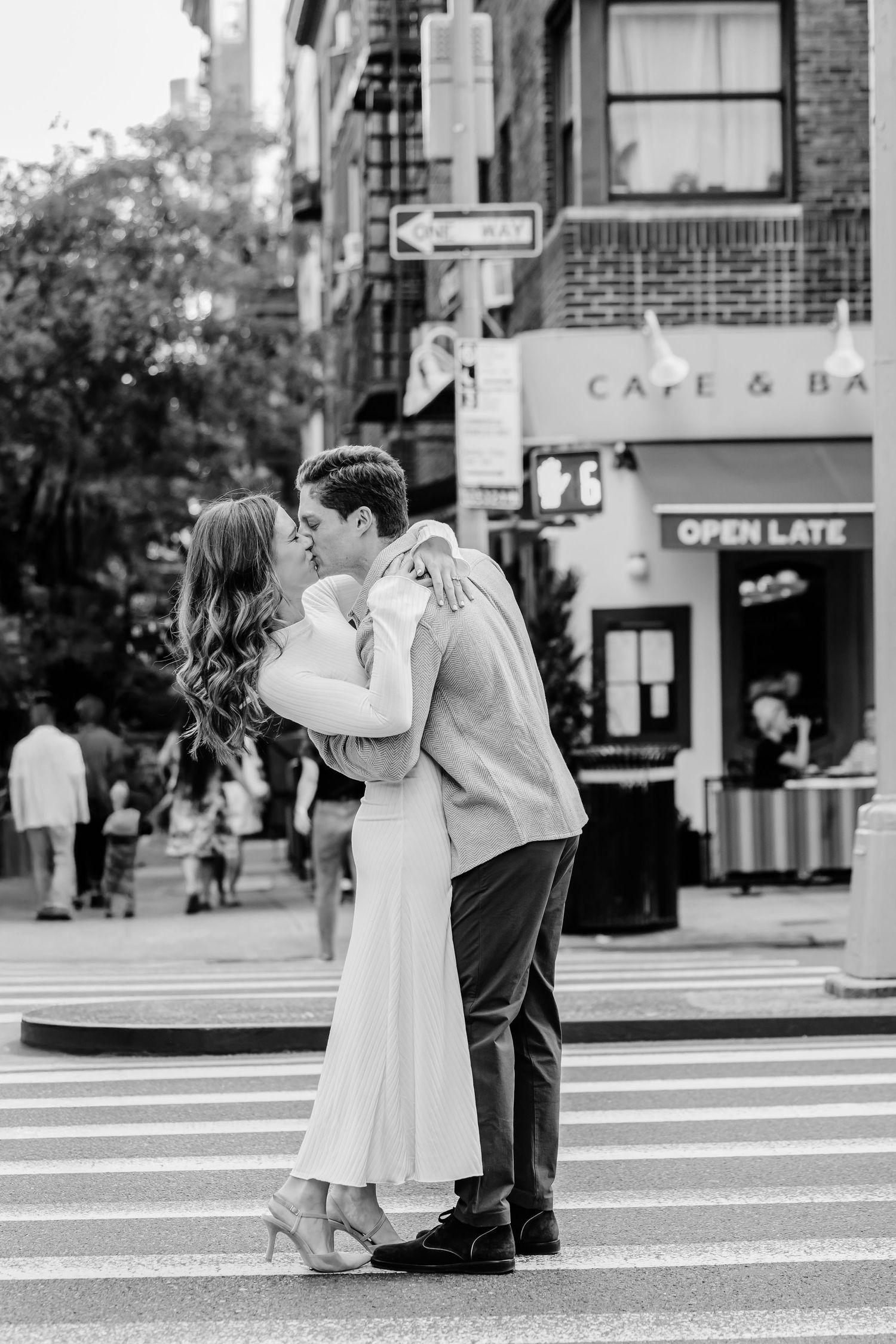 Black and white photo of couple kissing on a NYC crosswalk with cafe in background.