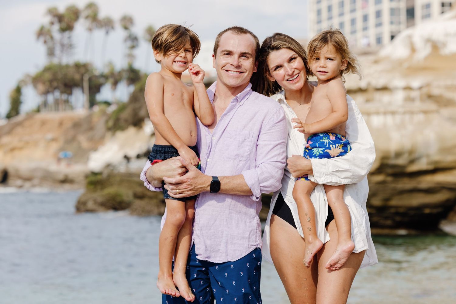 Happy family of four smiling together on a sunny beach with ocean and cliffs in background.