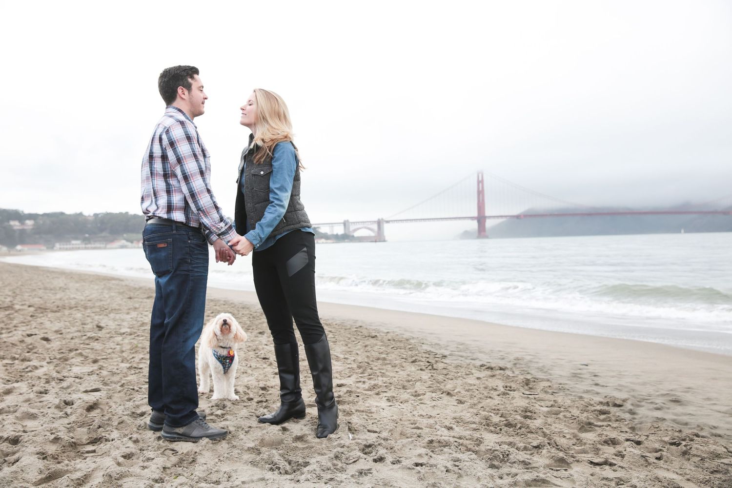 Couple holding hands on beach with Golden Gate Bridge in background and a pug dog.