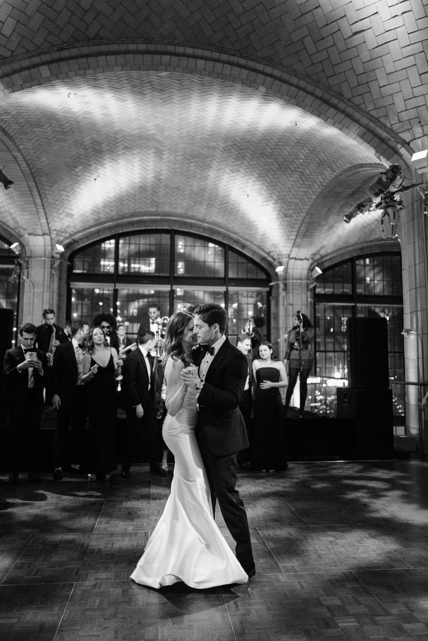 Bride and groom share an intimate first dance under vaulted brick arches surrounded by wedding guests.