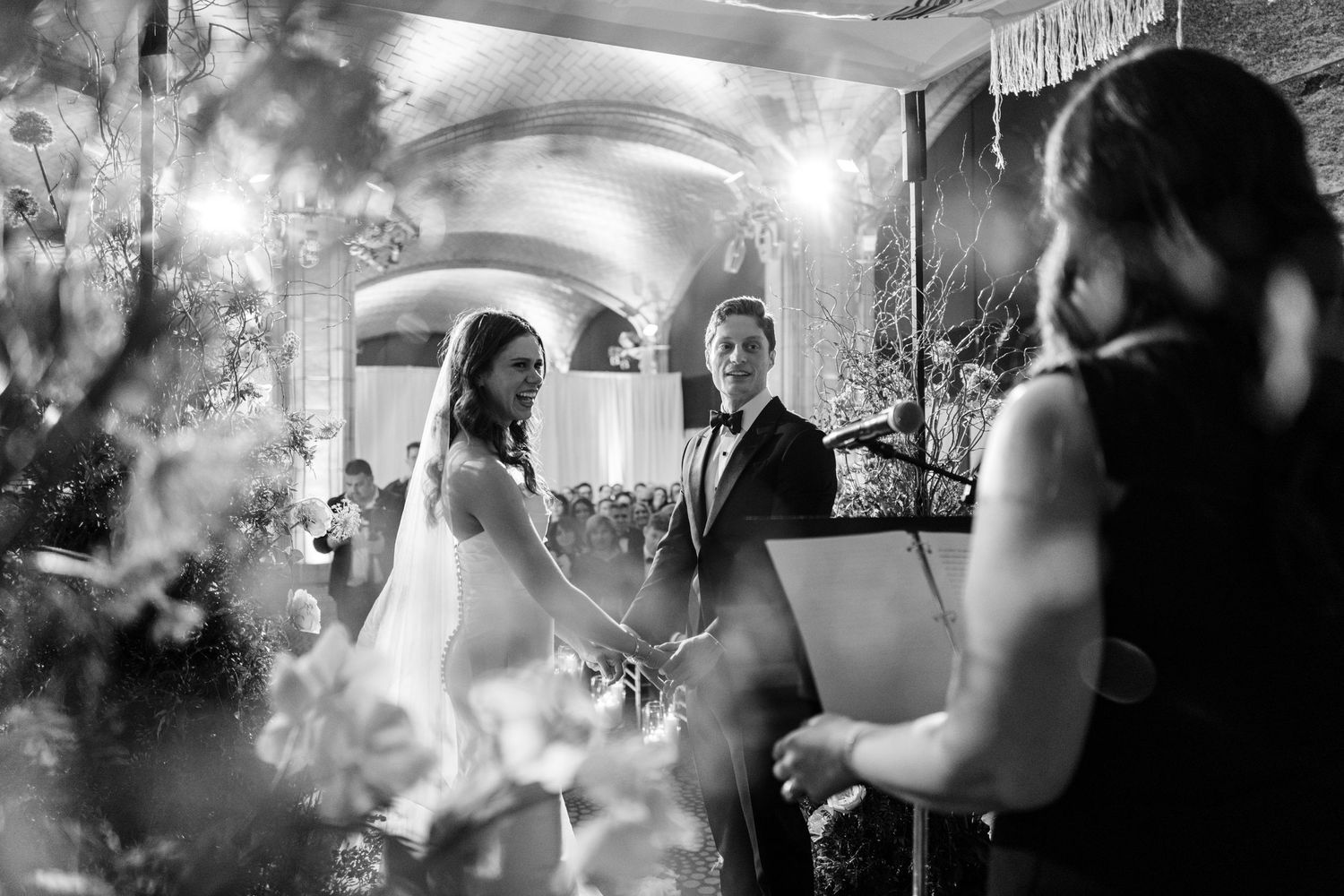 Black and white close-up of bride and groom holding hands at altar during intimate wedding ceremony.