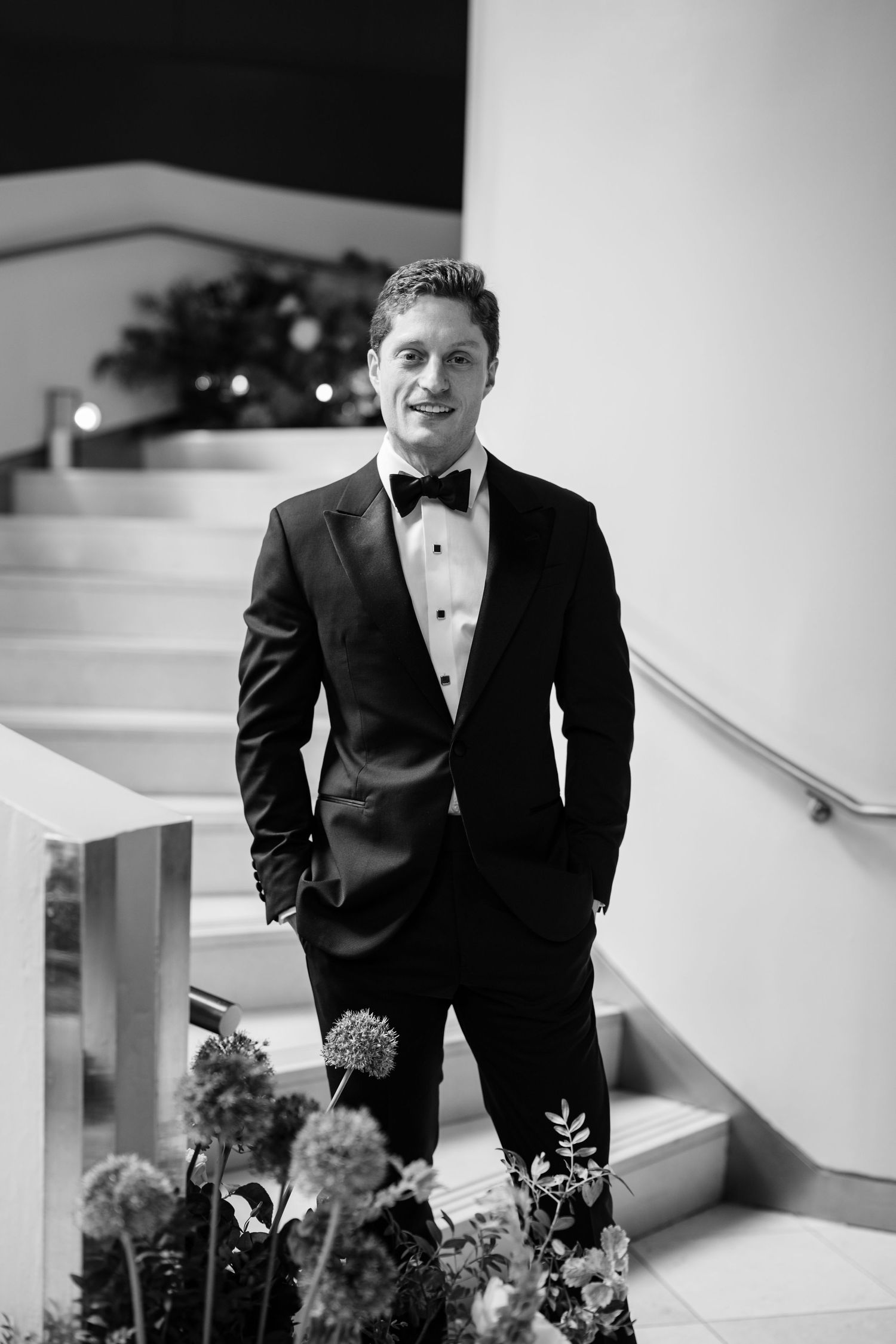 Black and white portrait of groom in tuxedo with bow tie standing on staircase with bokeh lights.