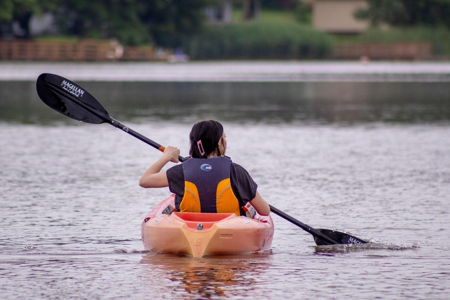 A kayaker in an orange life vest paddles a beige kayak on a peaceful lake.