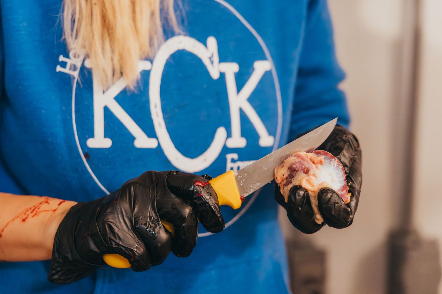Person wearing a blue KCK shirt and black gloves grips a glass beer bottle while working or serving drinks.