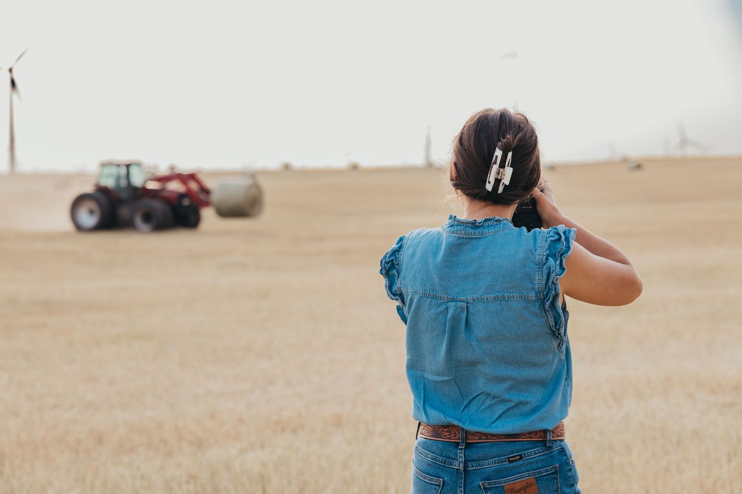 Person in blue denim looking out at a tractor working in an expansive dry field on a sunny day.