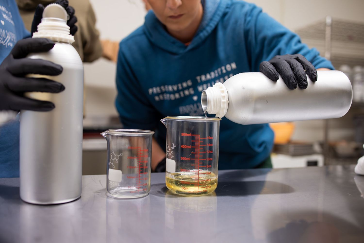 Laboratory worker pouring liquid ingredients into measuring beakers on steel table.