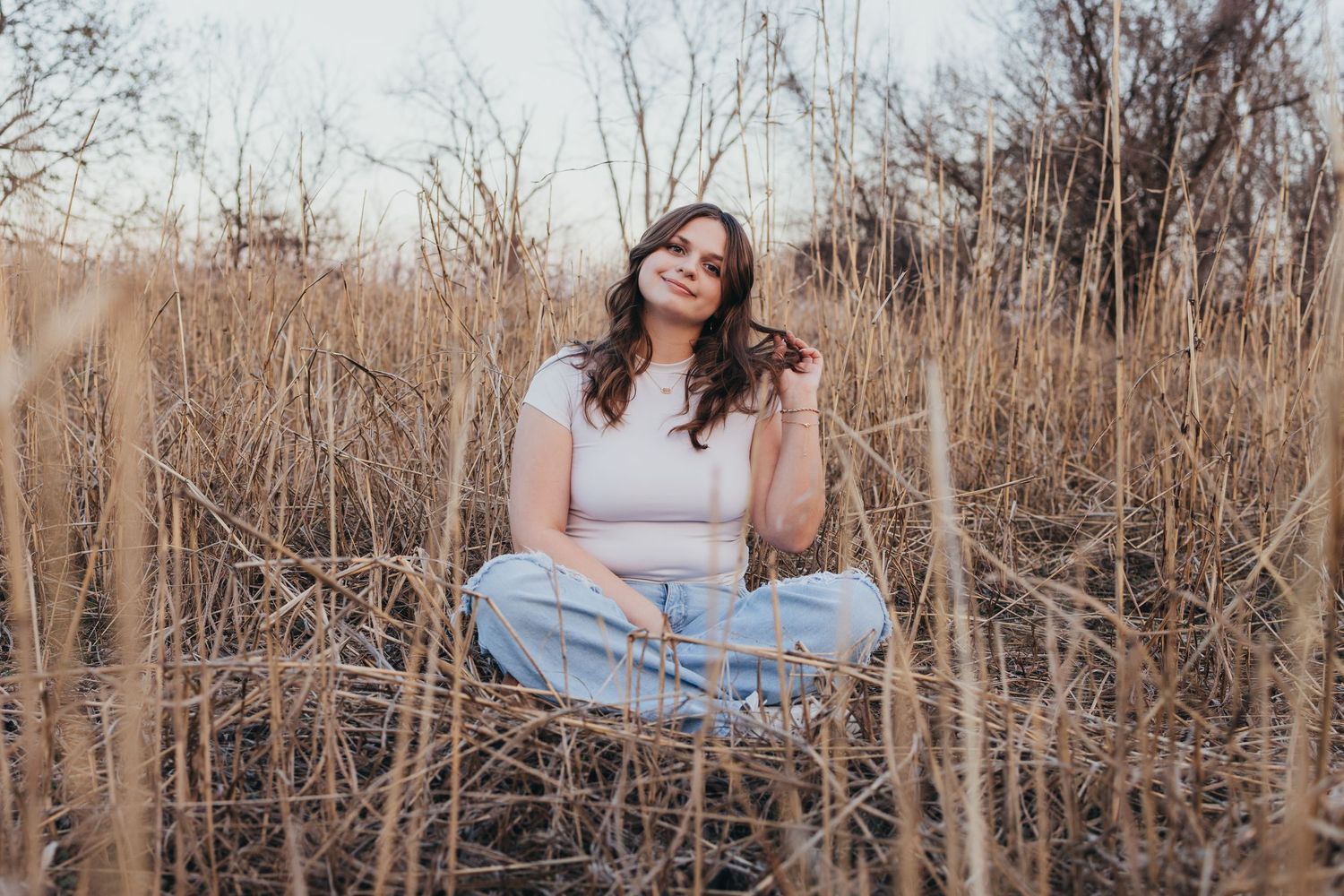 Young woman sitting cross-legged in dry golden grass field, wearing white top and jeans, smiling peacefully outdoors.