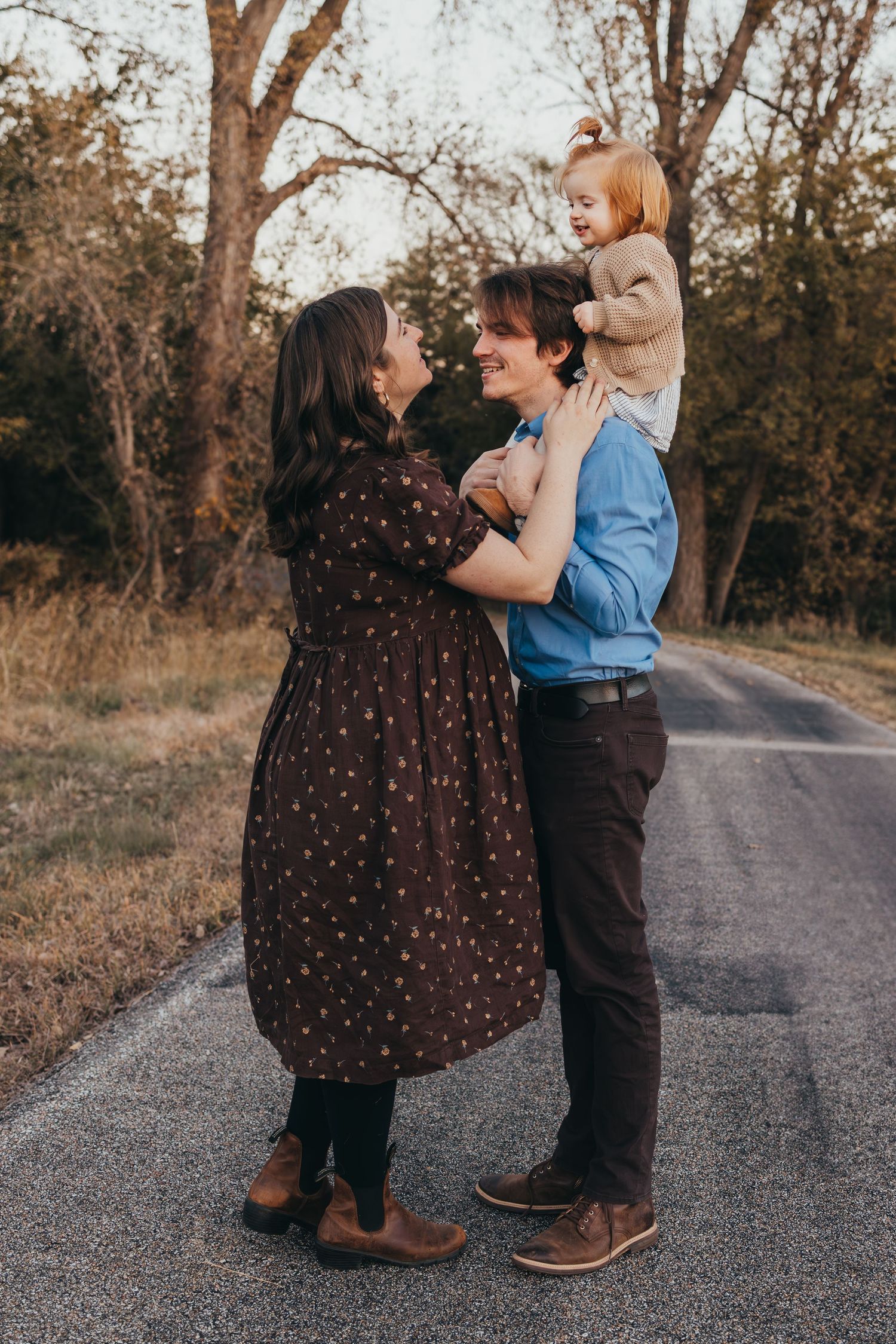 Family of three posing on a quiet road surrounded by autumn trees, parents smiling at each other while holding their toddler.
