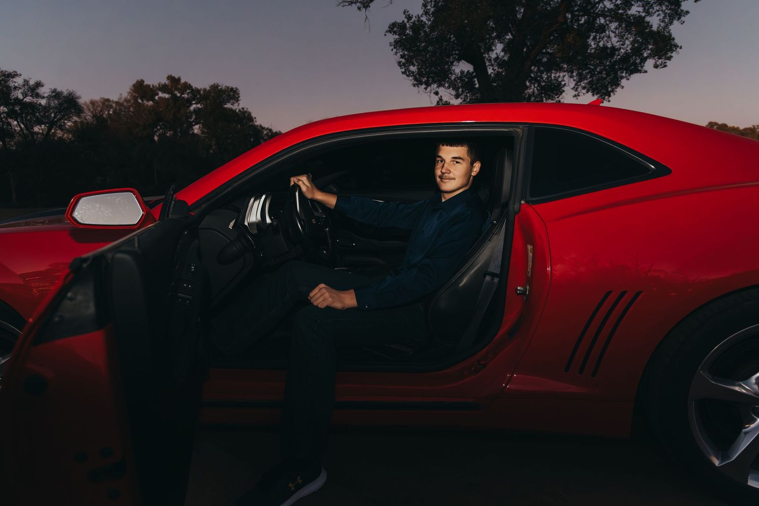 Young man sitting in red sports car at dusk, posing confidently in open door of sleek Camaro.