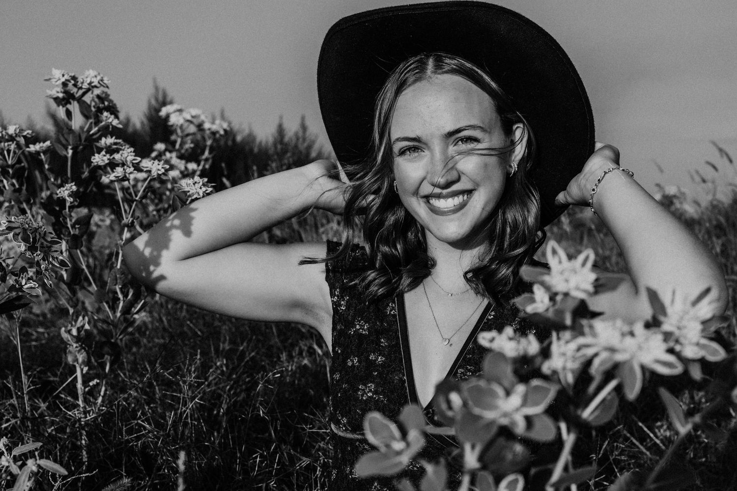 Black and white portrait of someone smiling joyfully in a hat among flowers in a garden.