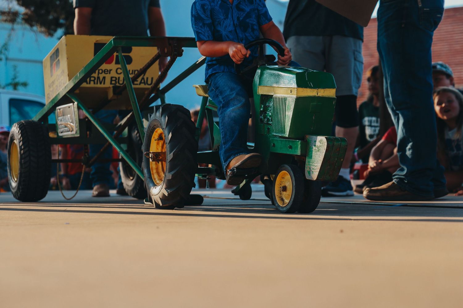 Small green toy pedal tractors lined up on a sidewalk during an outdoor event or gathering.