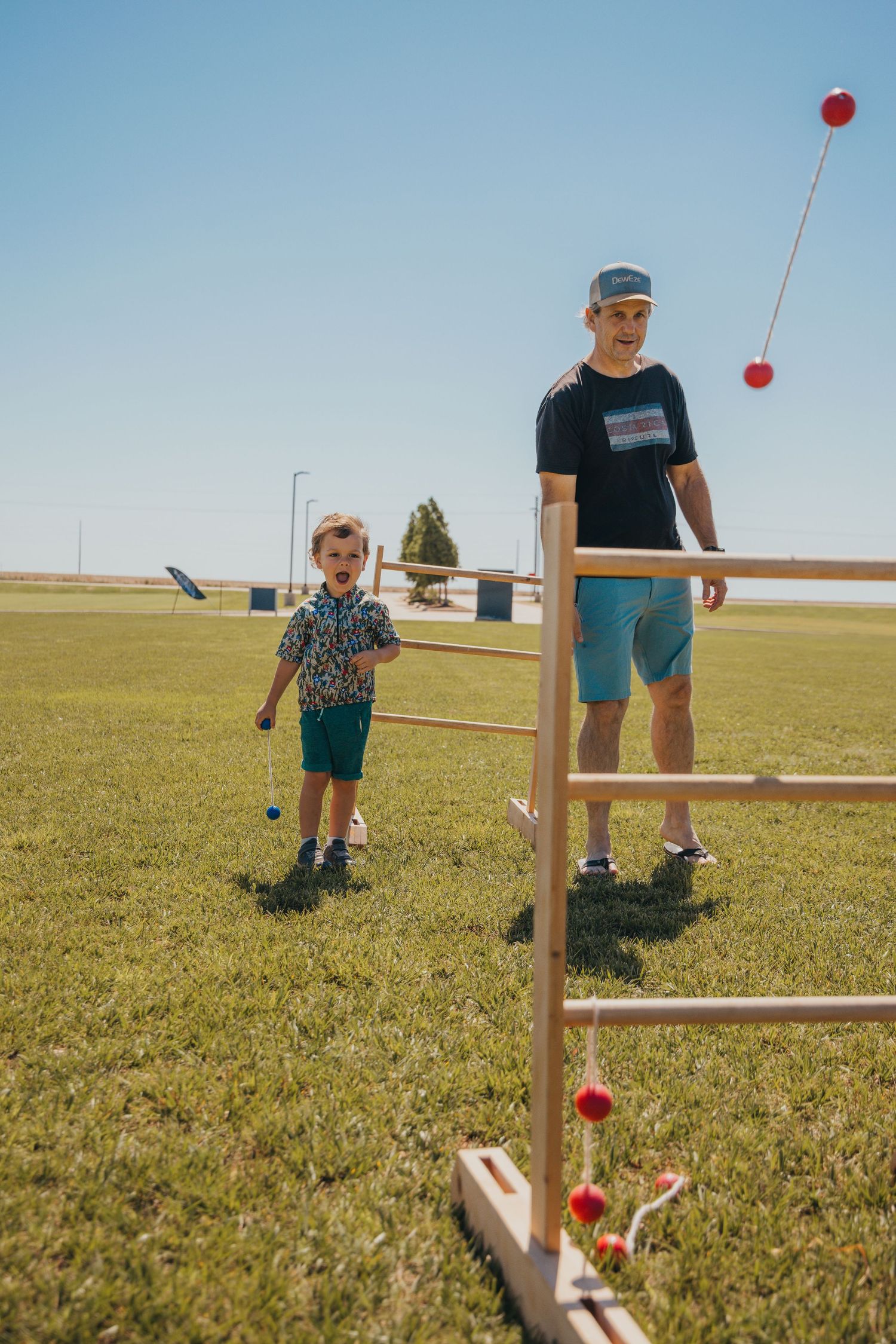 Two people play a ladder ball yard game with red bolas on a sunny day in a grassy field.