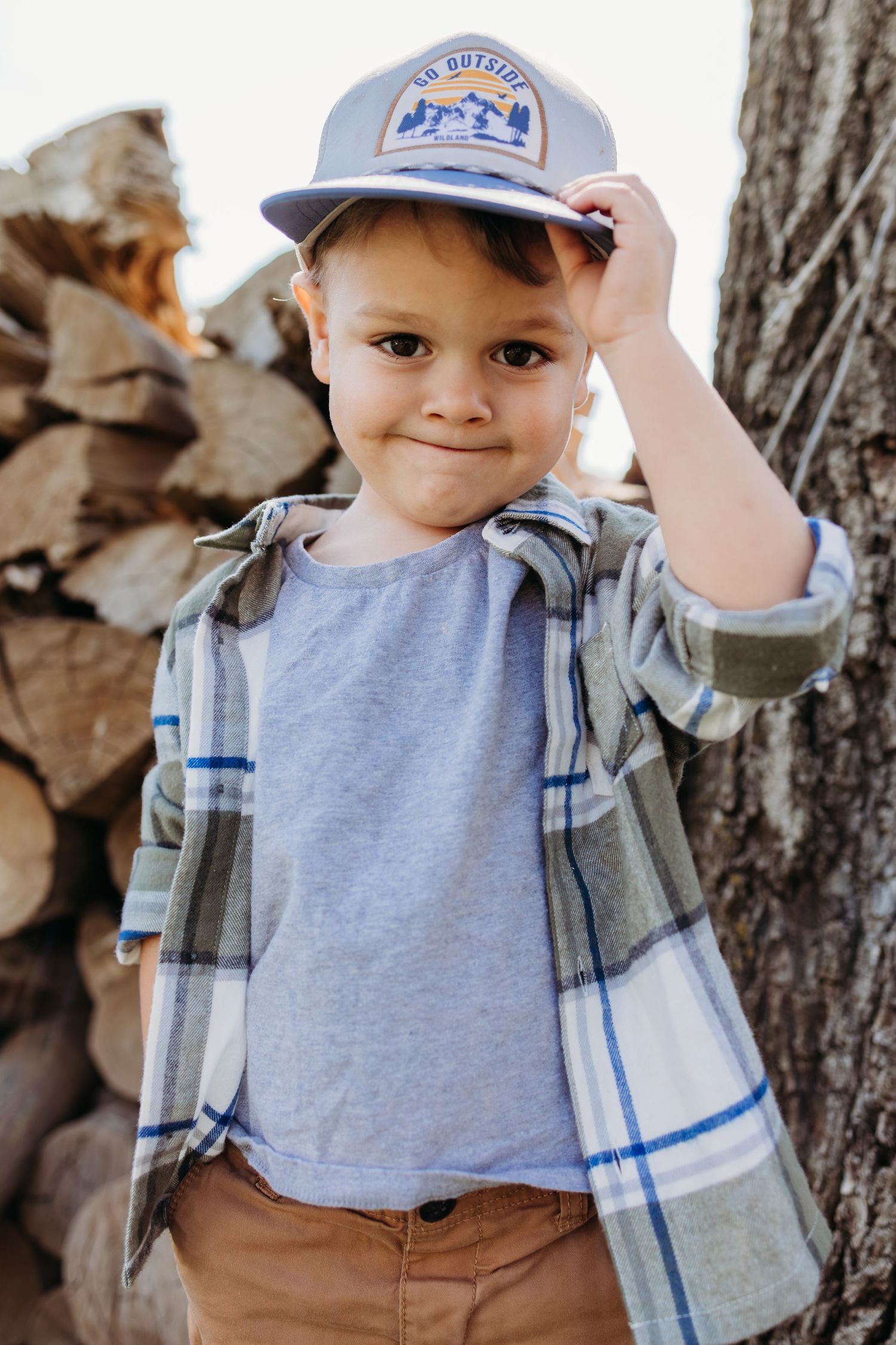 A child wearing a blue and white plaid shirt and baseball cap poses outdoors near a woodpile and tree.