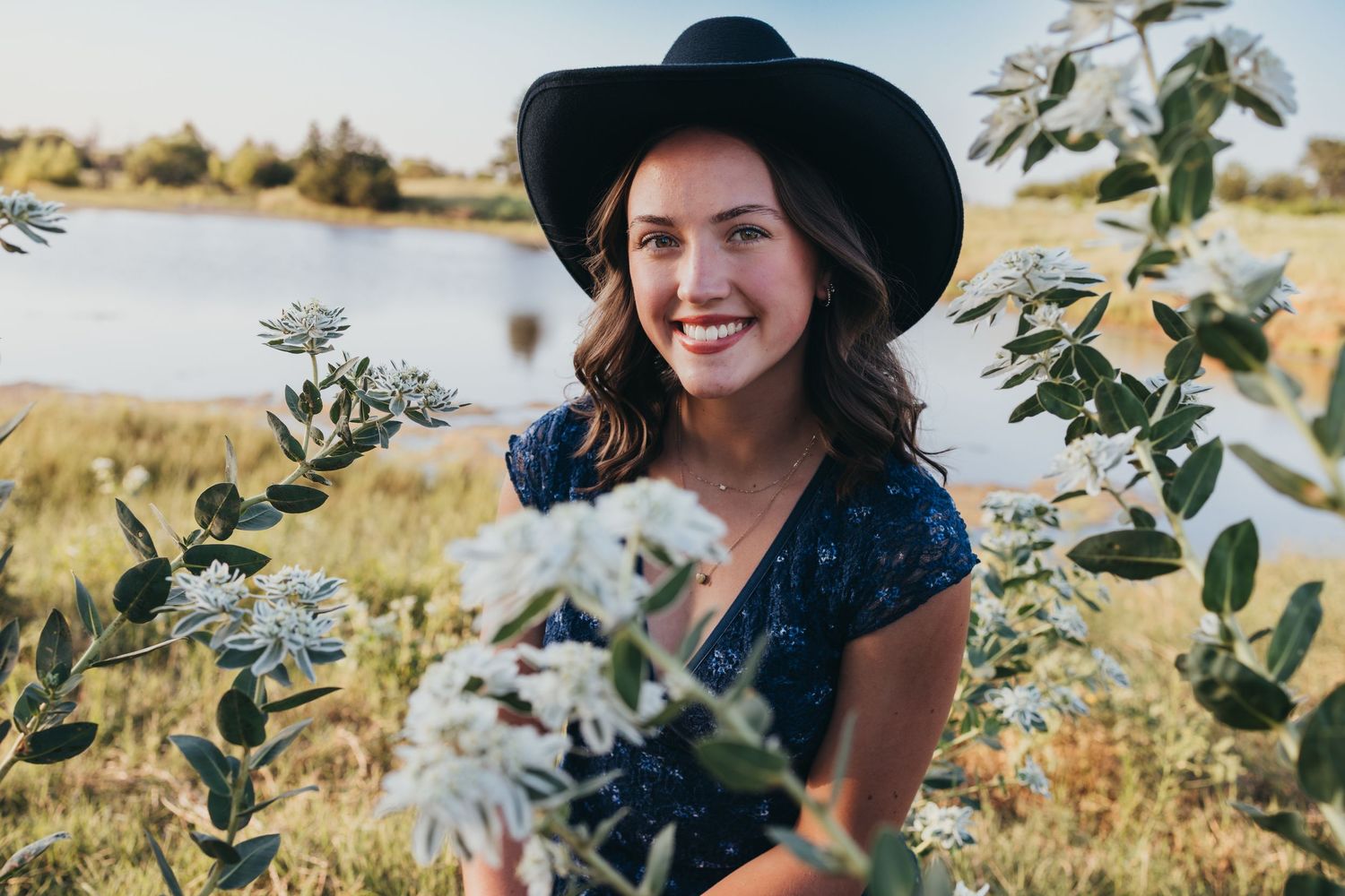Someone in a black hat poses happily in a field of white wildflowers on a sunny day.