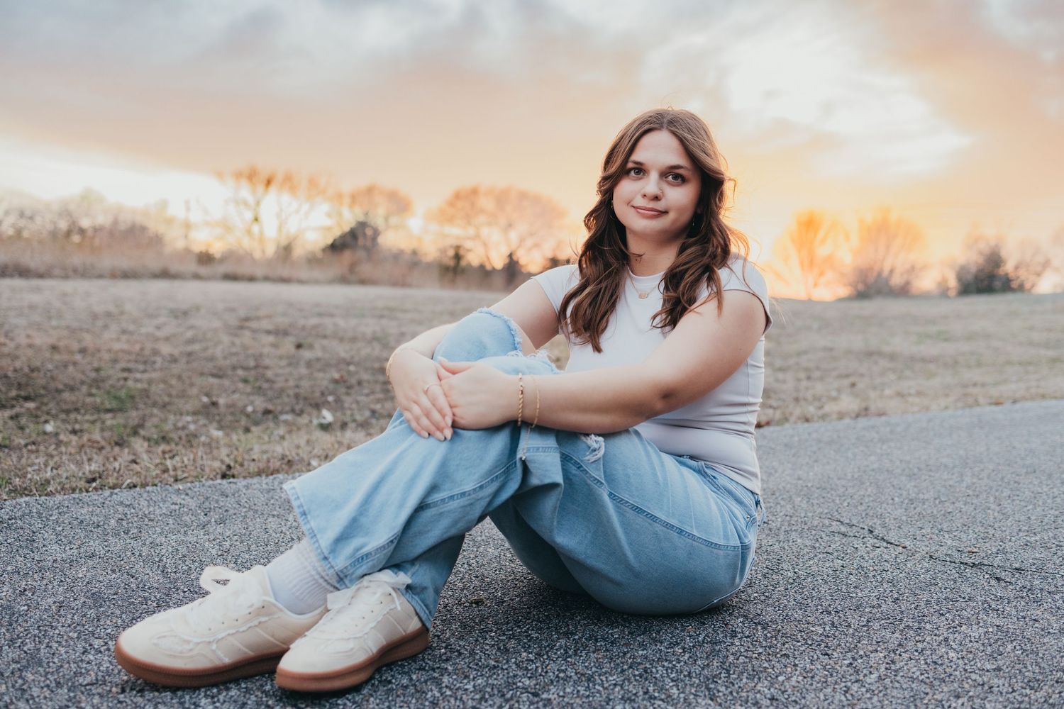 Young woman sitting on pavement at sunset, wearing jeans and white top, with golden sky and bare trees behind her.