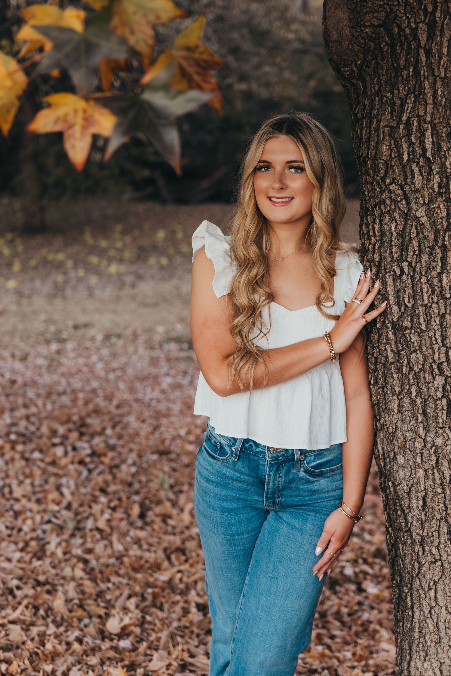 Young woman in white top and jeans leaning against a tree surrounded by autumn leaves in a park.