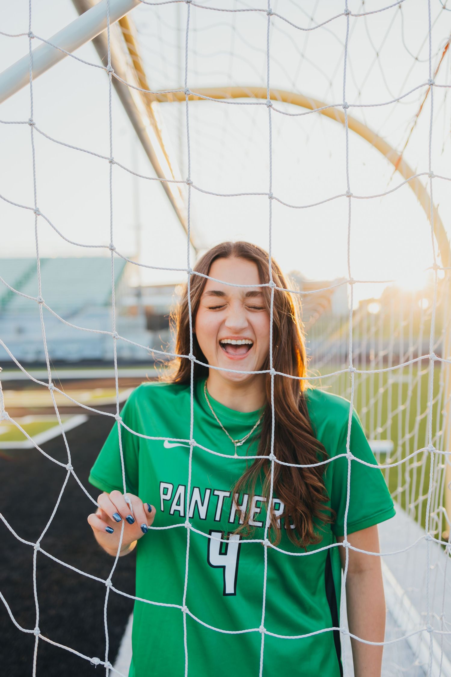 A smiling girl in a green Panther #4 soccer jersey poses behind a goal net on a soccer field.