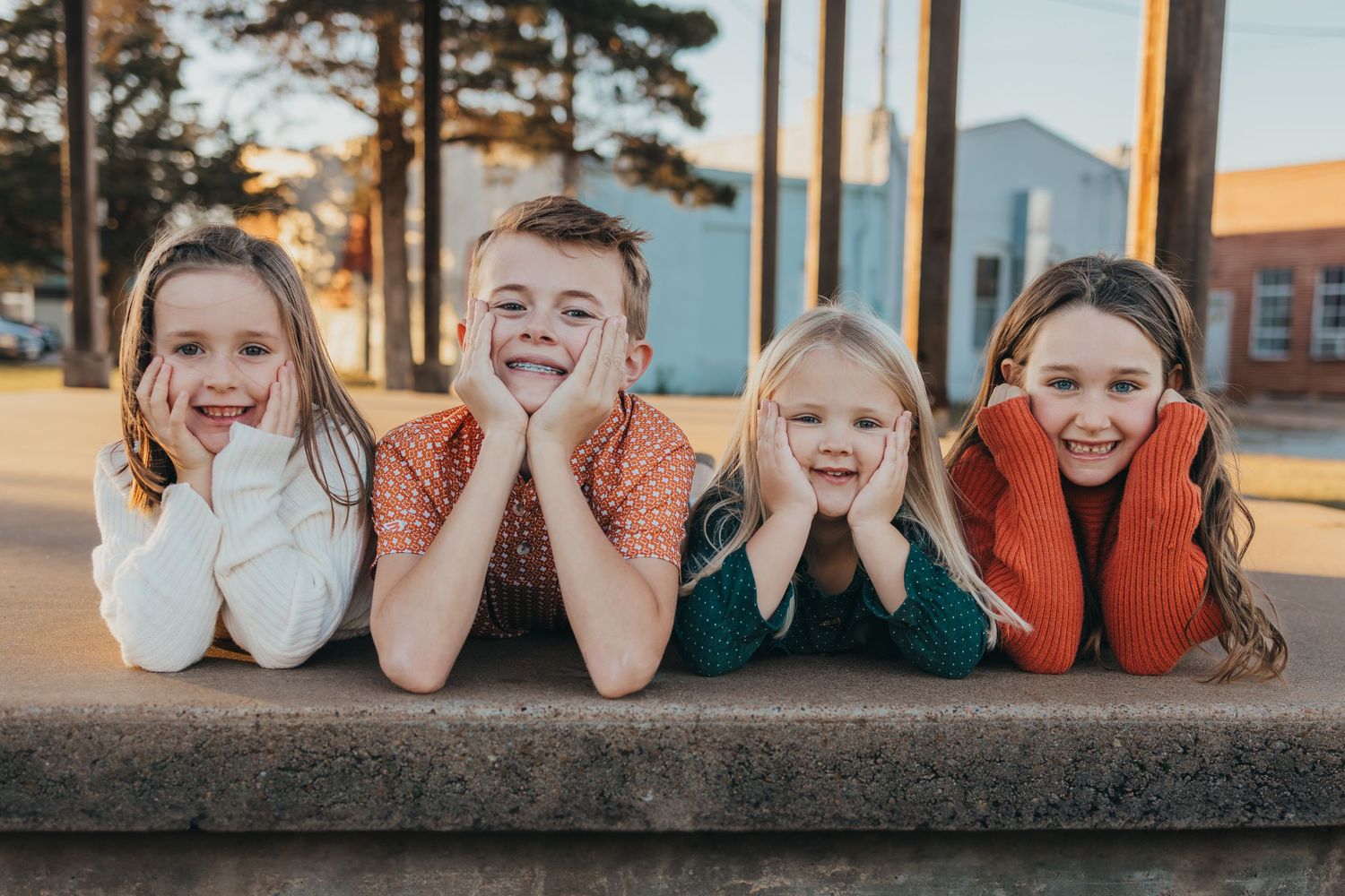 A row of children lying on their stomachs with hands cupped around their faces in an outdoor setting.