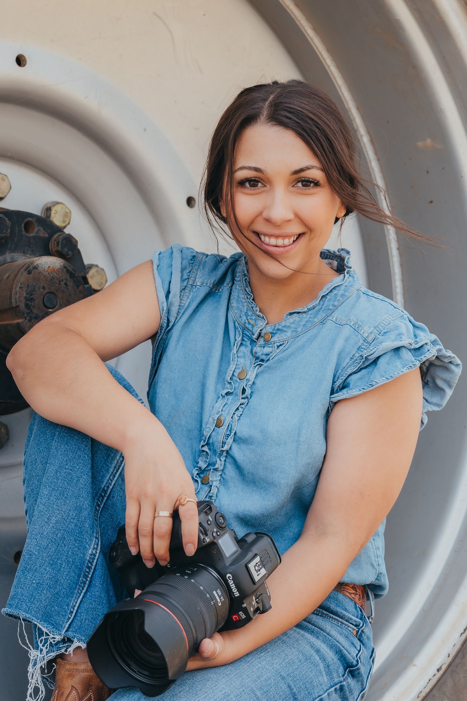 Professional photographer wearing denim outfit sits with camera gear against industrial backdrop.