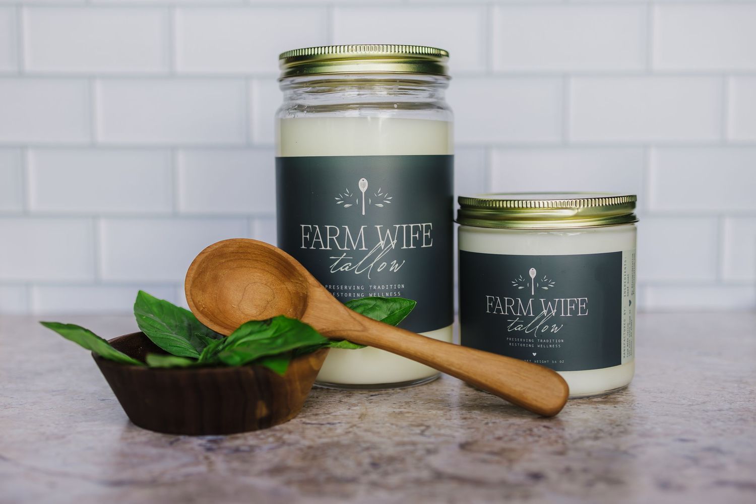 Two Farm Wife candles in dark green jars displayed with fresh herbs and a wooden spoon on a marble countertop.