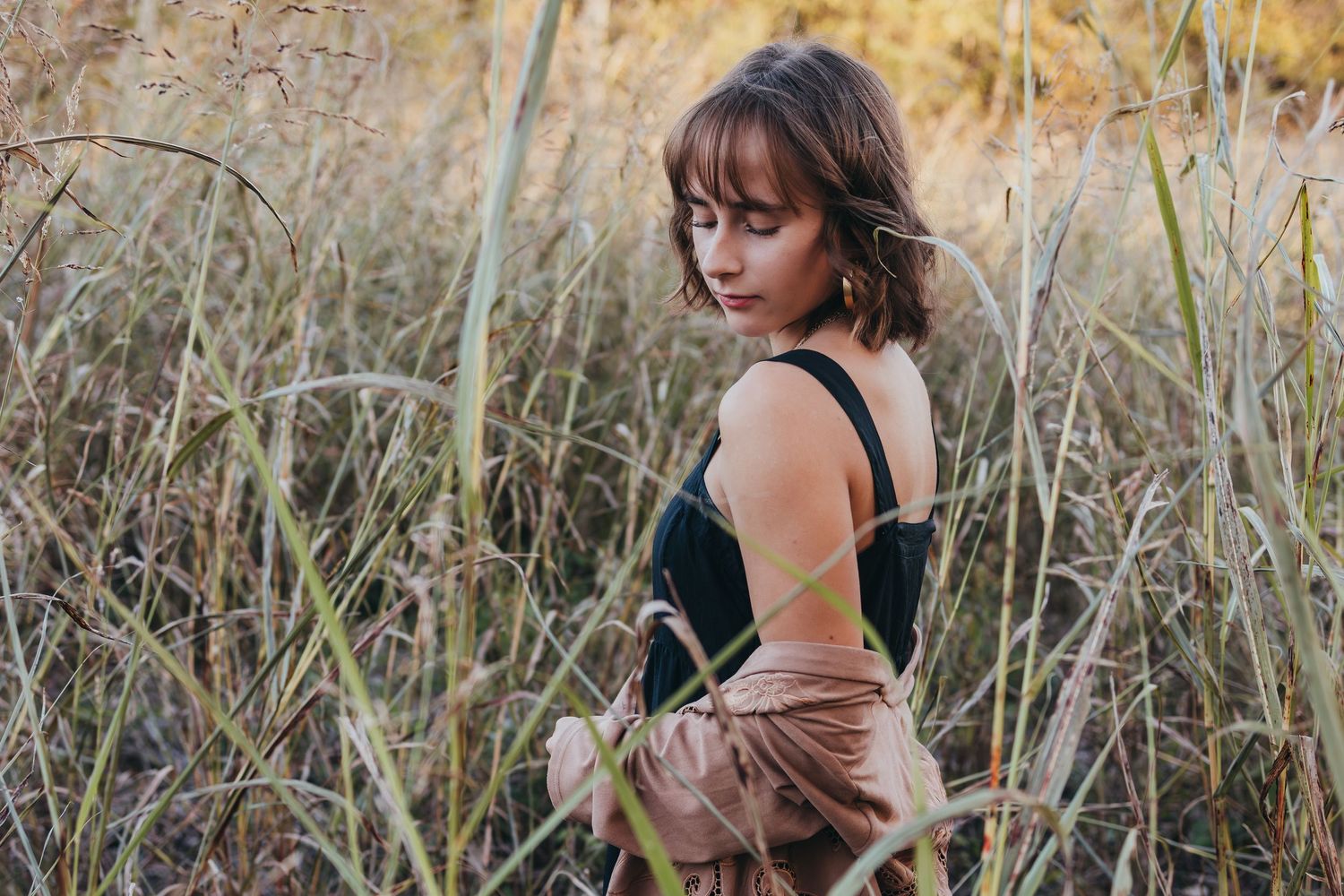 A young woman in a black top and pink skirt stands among tall golden grasses in a serene outdoor field setting.
