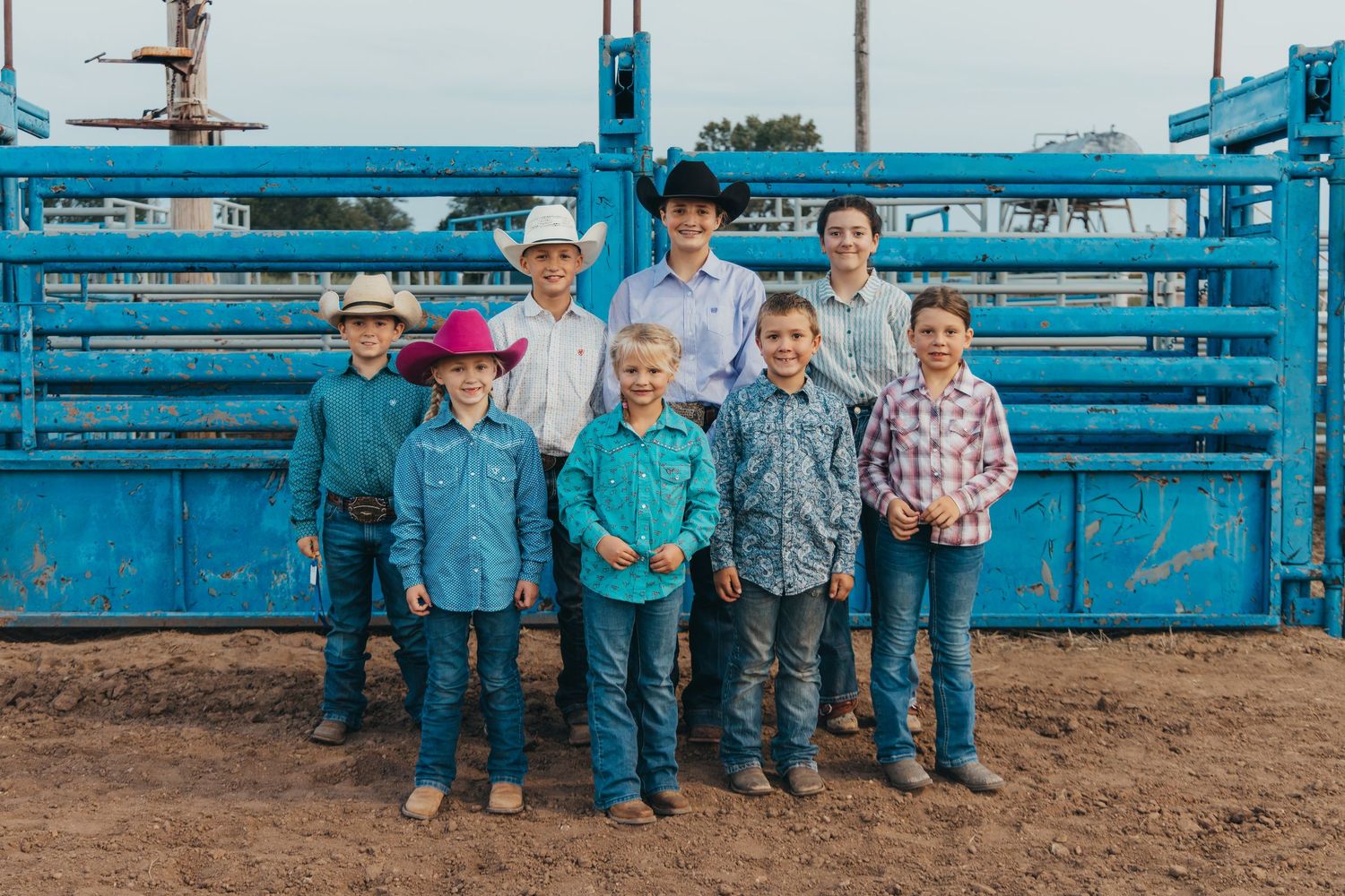 Group of young children standing in front of bright blue livestock gates at a rustic ranch setting.