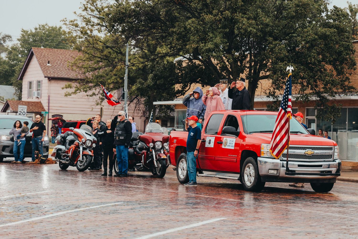 Red pickup truck and motorcycles lined up for parade on rainy street.