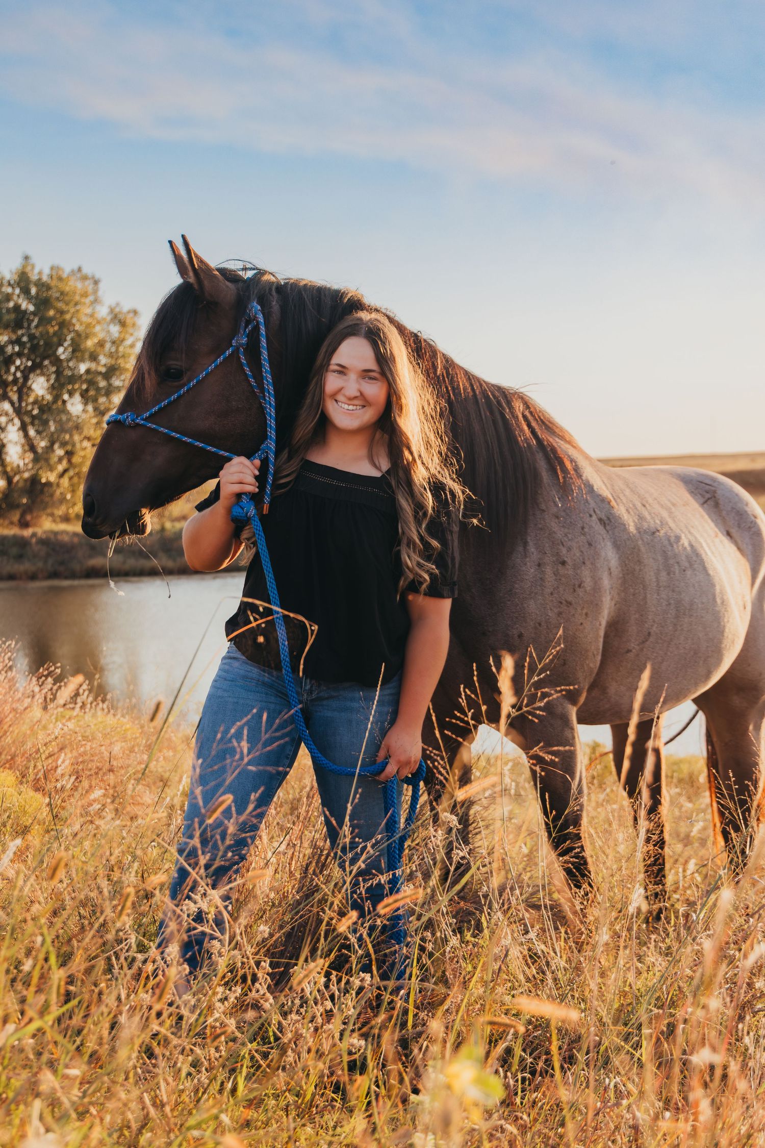 Young woman smiling and holding a blue rope lead while standing beside a roan horse in a golden field.