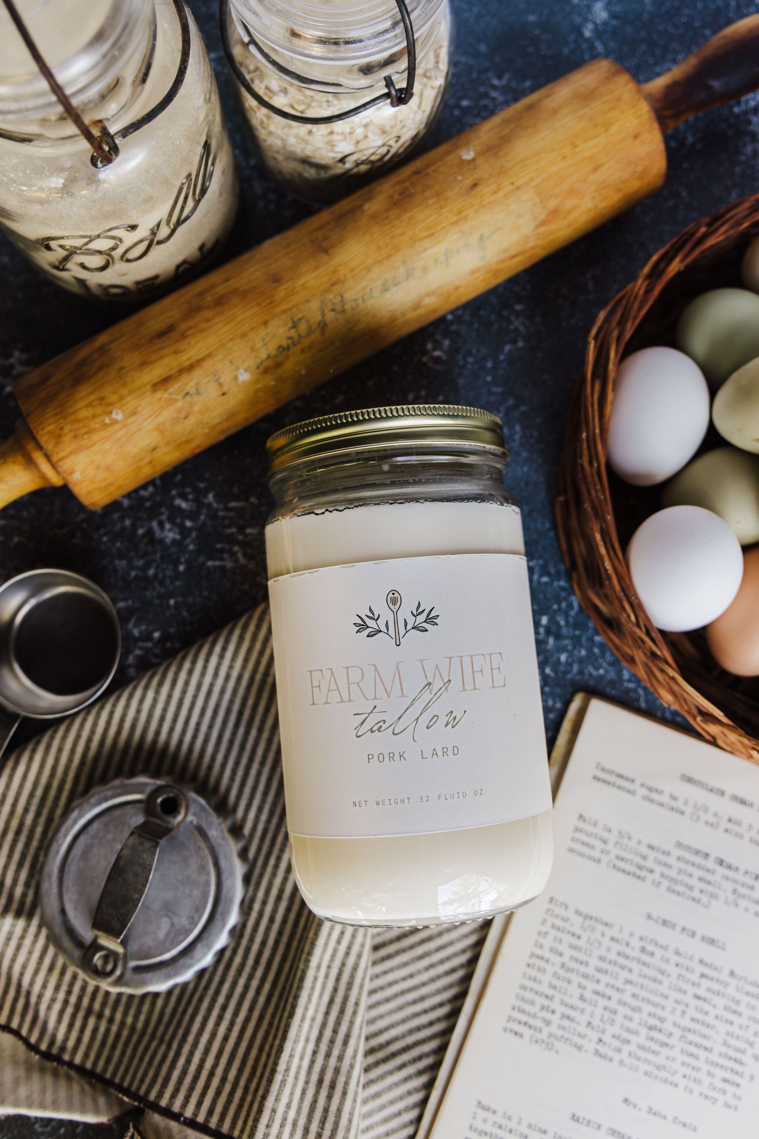 A jar of Farm Wife Tallow Pork Lard sits on a dark surface surrounded by eggs, a rolling pin, and mason jars.