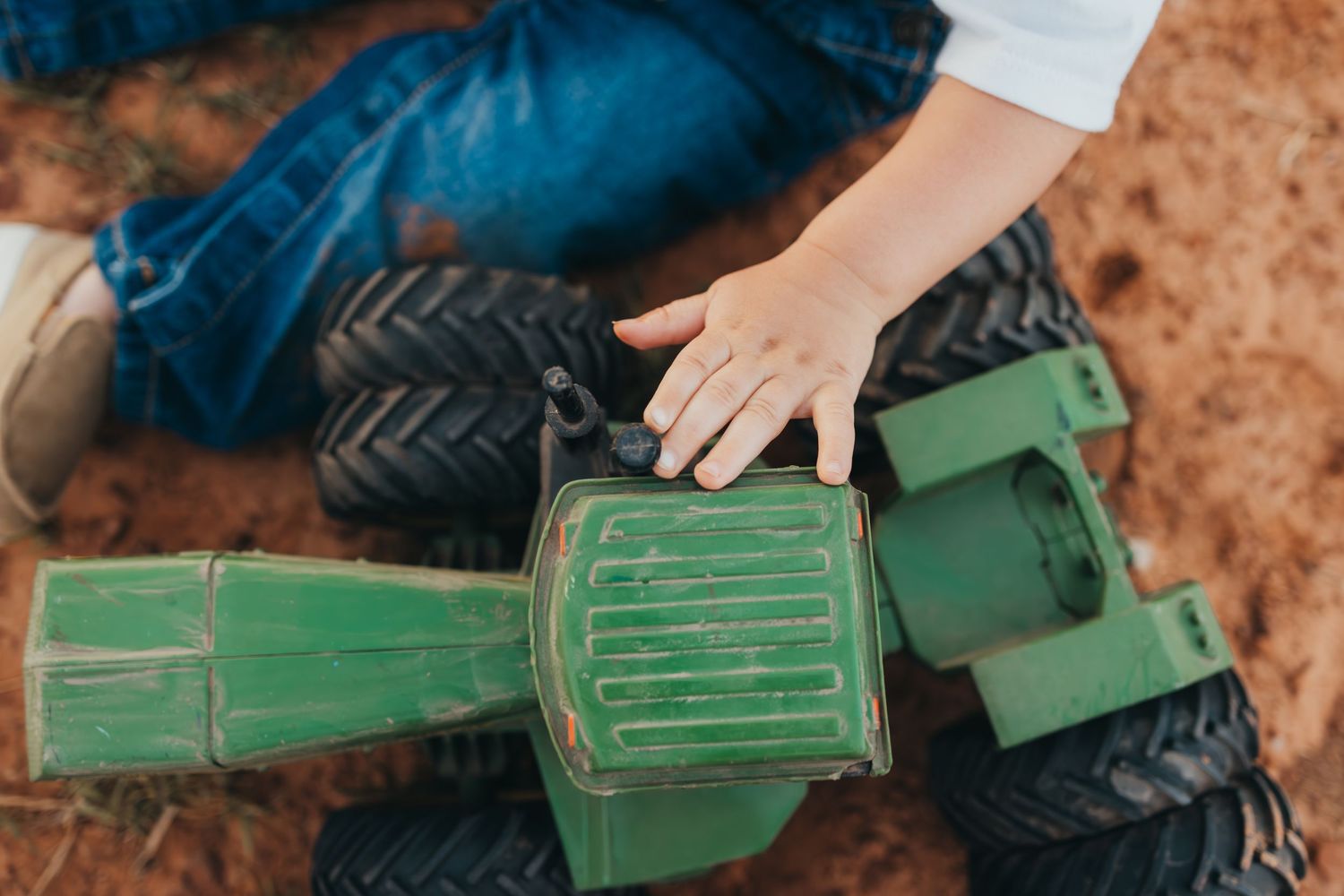 Hand touching green toy tractor parts on red dirt surface.