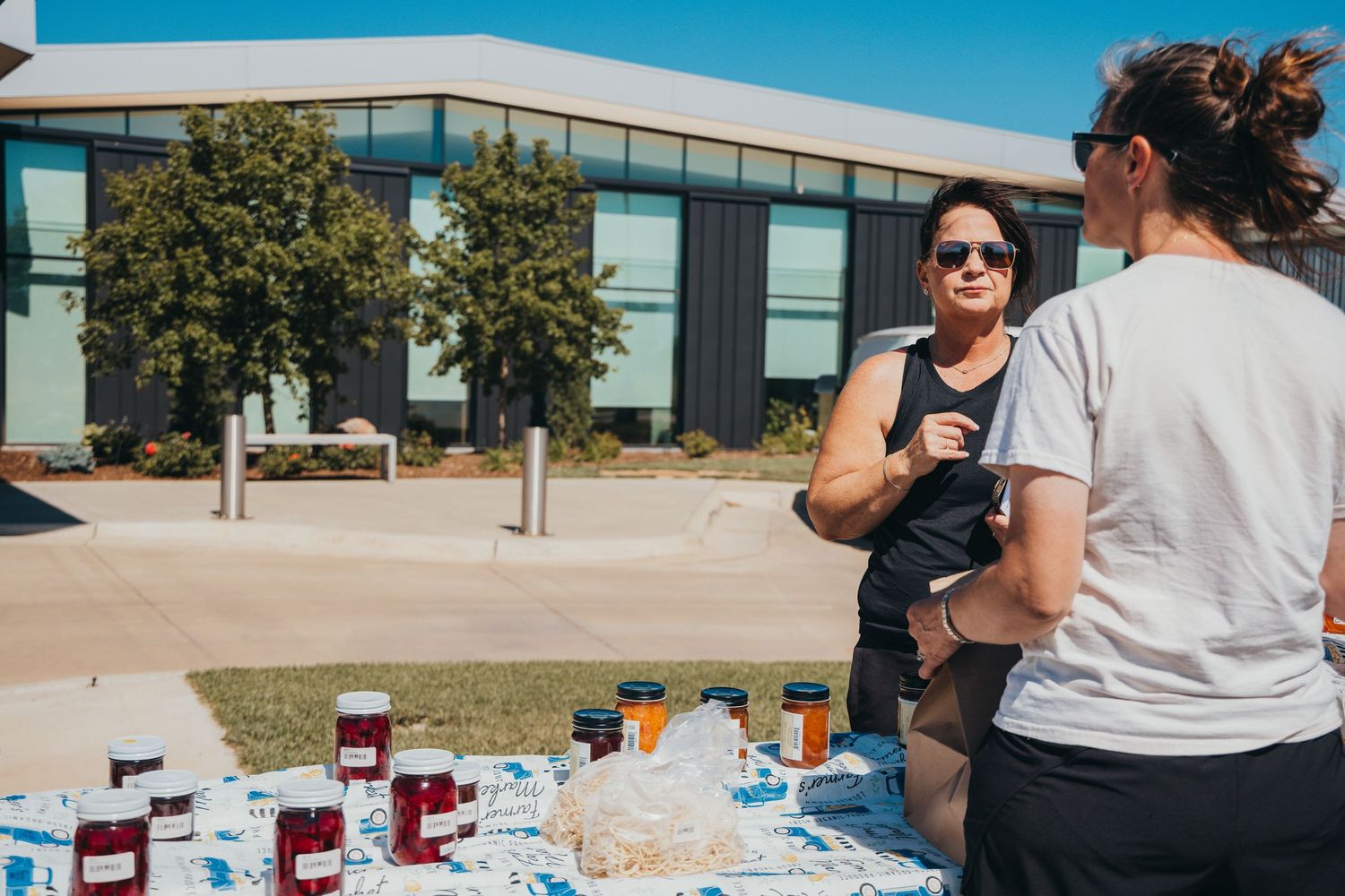 People chatting at an outdoor event with beverages and supplies spread out on a table under bright blue sky.