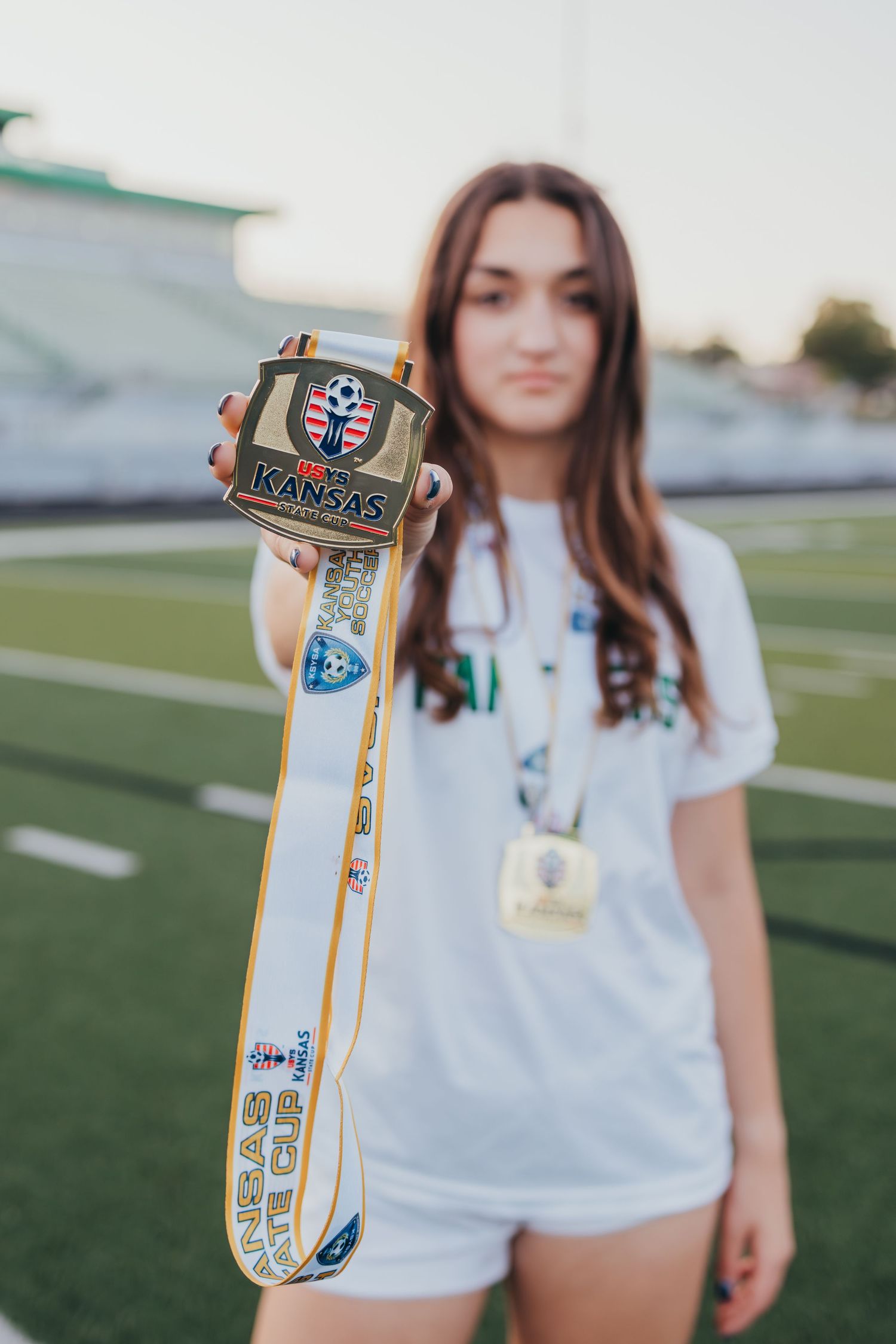 Young female soccer player holding a Kansas state championship medal and ribbon on a green athletic field.