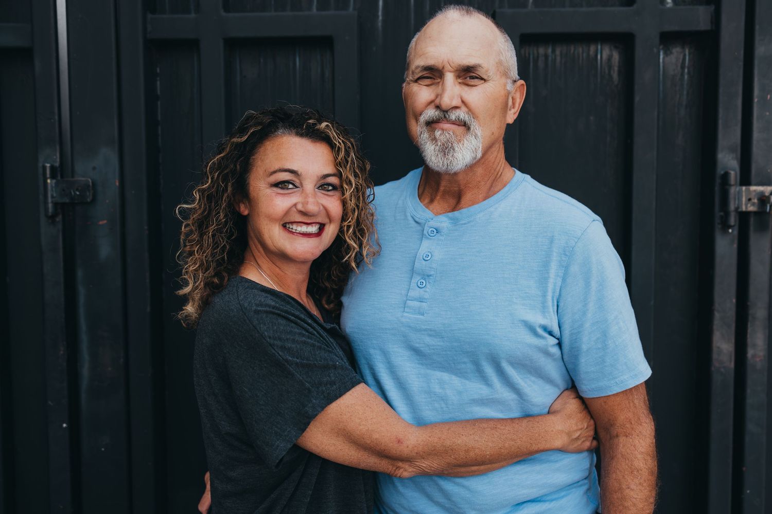 A casual outdoor portrait of two people wearing blue and black shirts against a dark wall.