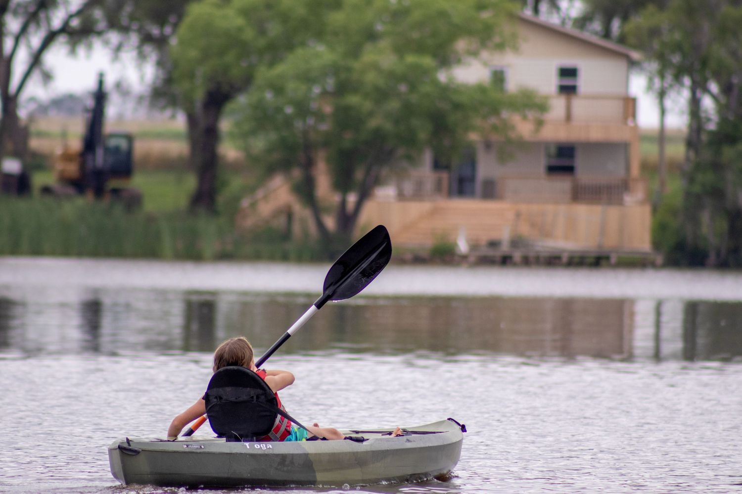 A lone kayaker paddles a light-colored kayak on a serene lake with buildings in the background.