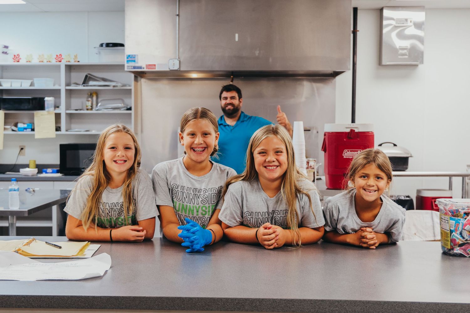 Students and instructor gather in a school kitchen classroom with blue supplies on the counter.