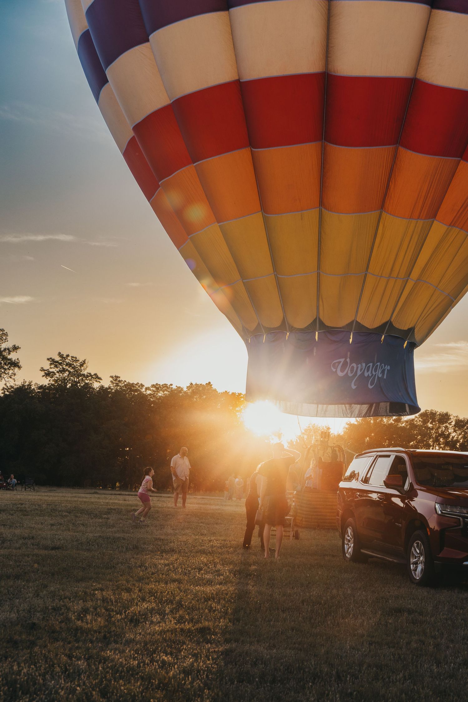 Hot air balloon silhouetted against dramatic sunset with spectators and cars in foreground.