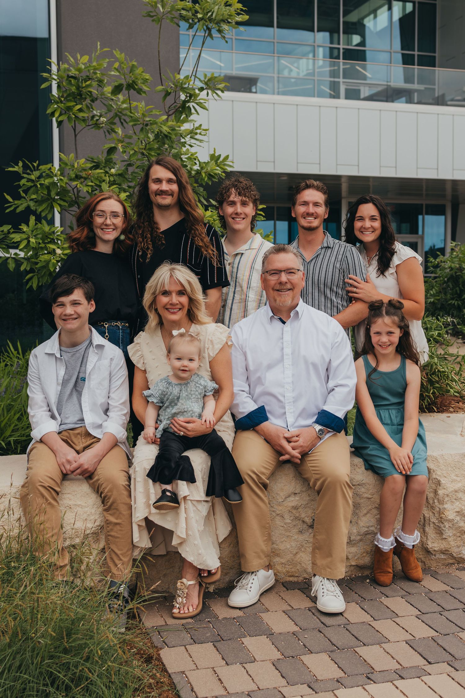 Multi-generational family sits together on brick steps in front of modern office building with greenery.