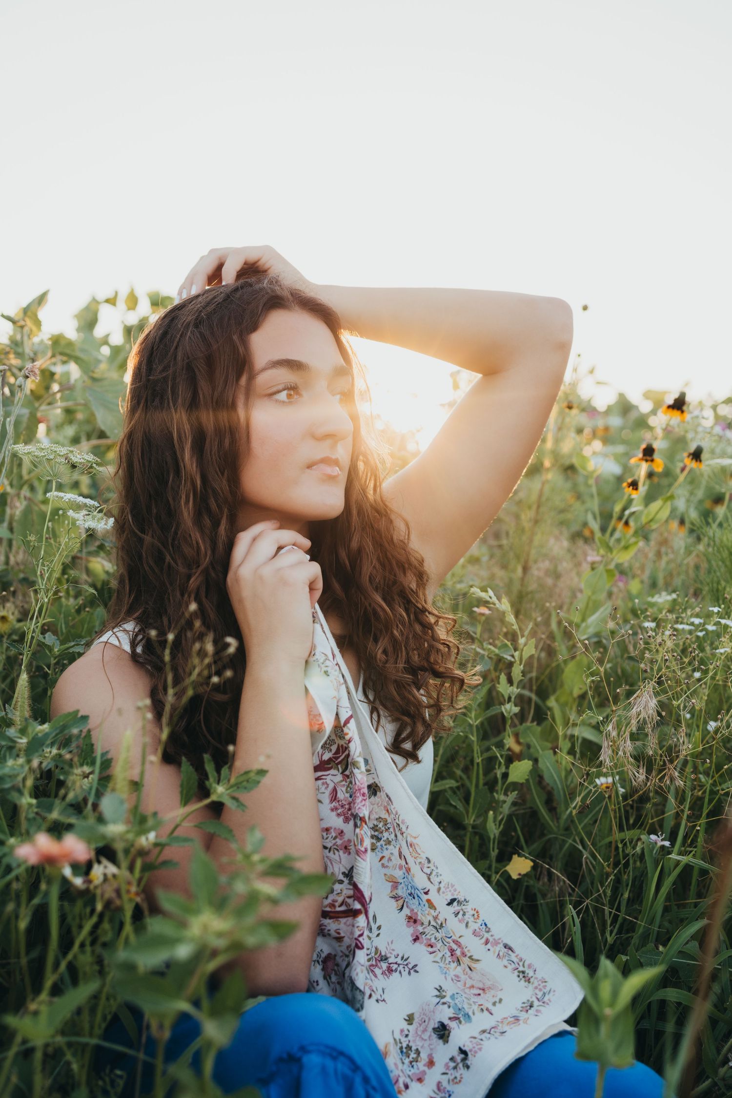 Person in a floral top sits dreamily among wildflowers at sunset in a summer meadow.