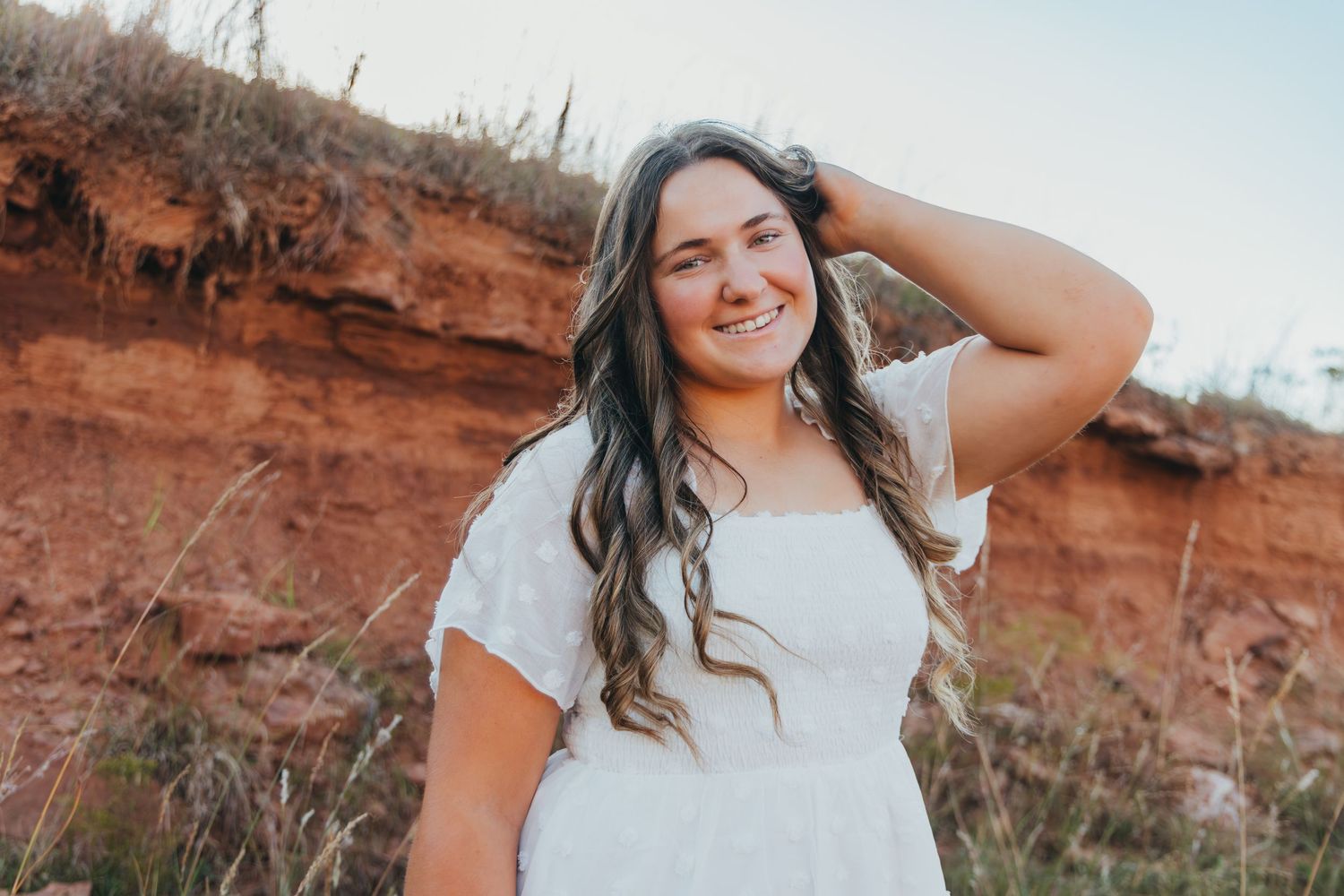 A woman in a white dress poses against red rock formations in a desert landscape at sunset.