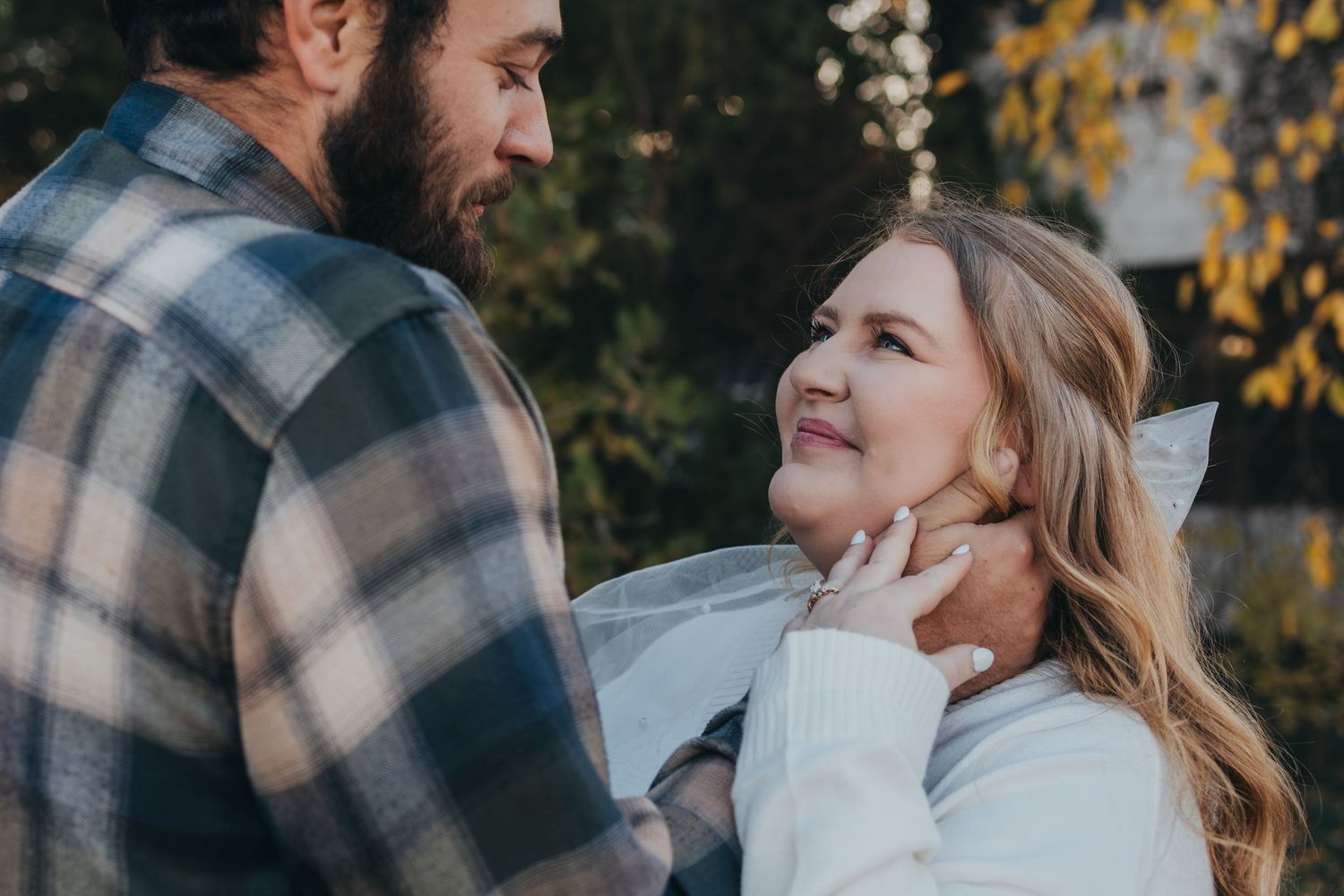 A couple shares a romantic moment outdoors in autumn, man in plaid shirt gently holding womans face as she smiles up at him.