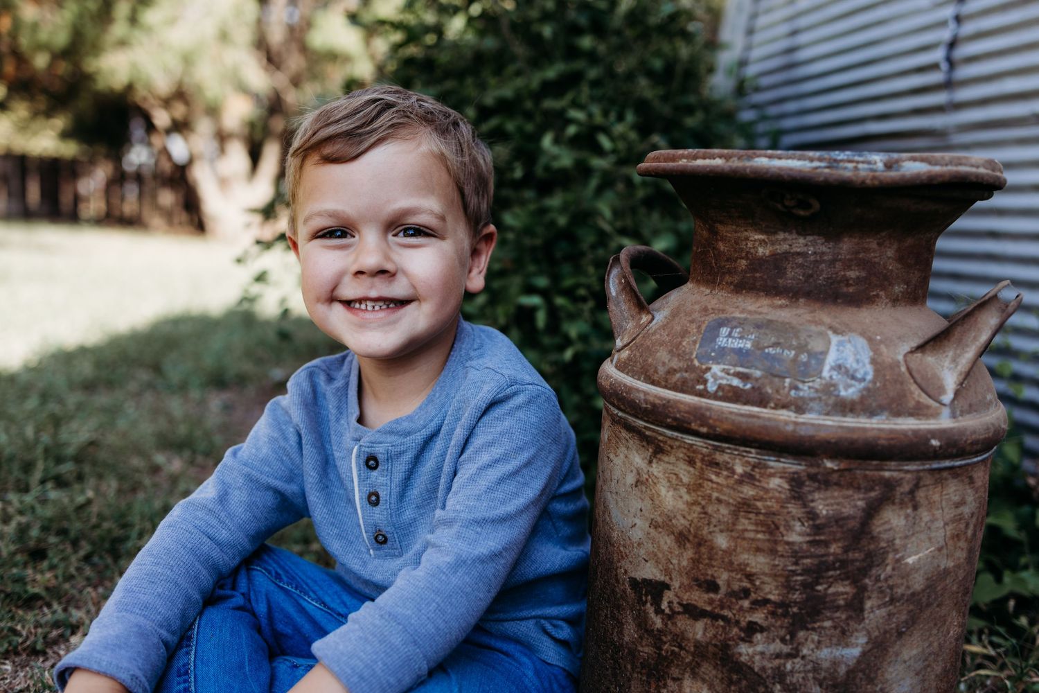 A child in a blue henley shirt sits outdoors next to a rustic metal milk can on a sunny day.