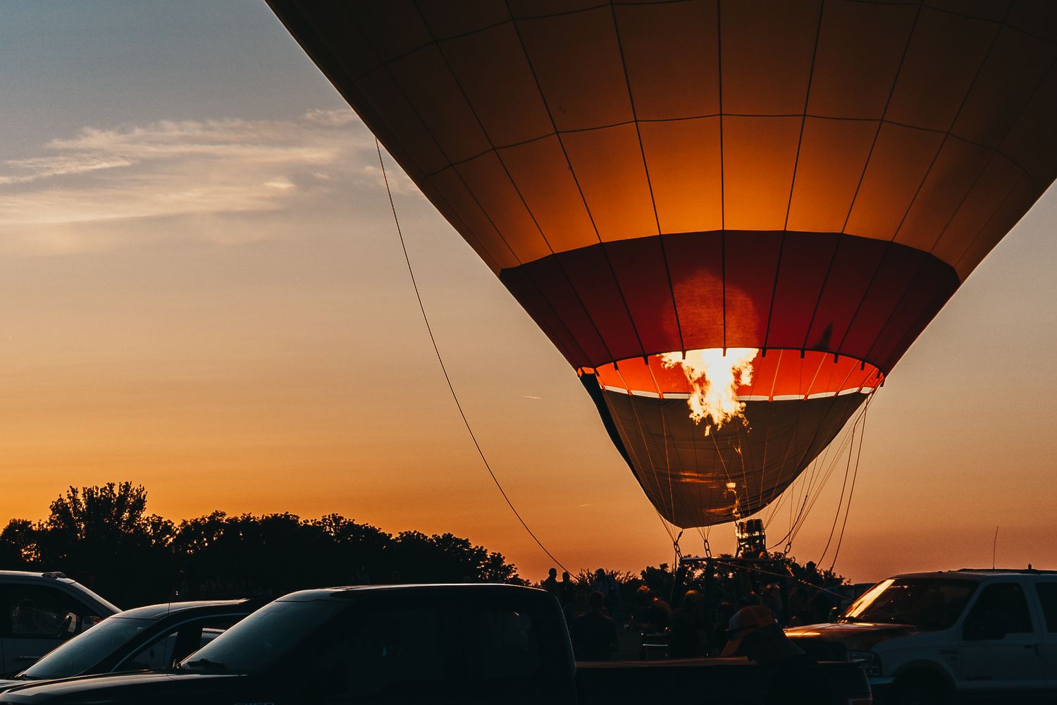 Hot air balloon glowing orange against sunset sky with cars silhouetted below.