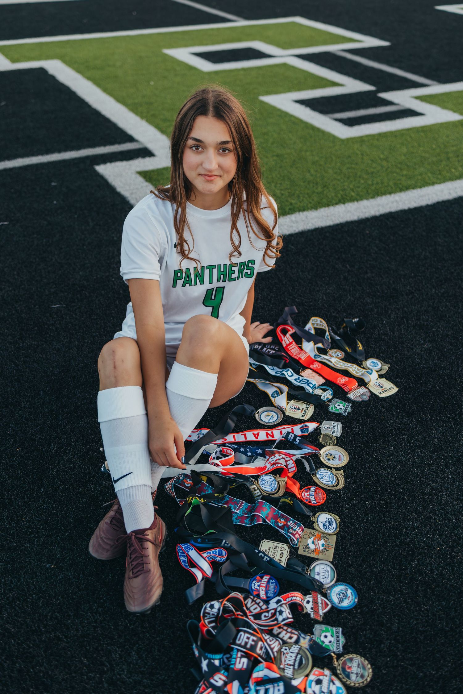Female soccer player in Panthers #4 jersey sits on turf field surrounded by a collection of medals and awards.